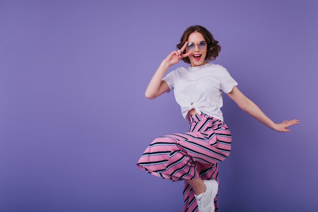 Ragazza bianca vaga in scarpe da ginnastica alla moda che salta sulla parete viola. Debonair donna dai capelli corti con occhiali da sole che scherza durante il servizio fotografico.