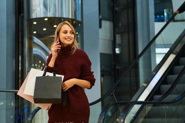 Ragazza bionda abbastanza allegra in maglione lavorato a maglia felicemente parlando al cellulare nel moderno centro commerciale