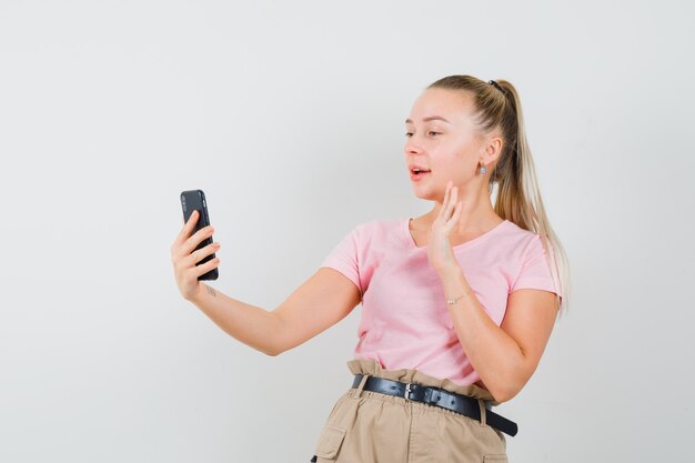 Ragazza bionda agitando la mano in chat video in t-shirt, pantaloni e guardando gioviale, vista frontale.