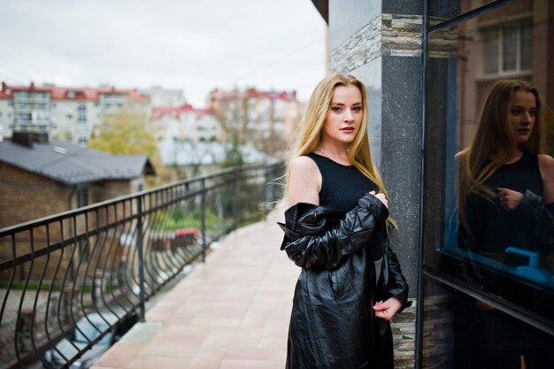 Ragazza bionda alla moda con un lungo cappotto di pelle nera posata contro la grande finestra dell'edificio