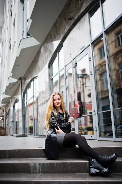 Ragazza bionda alla moda in lungo cappotto di pelle nera posata alle scale