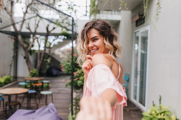 Ragazza bionda allegra che osserva sopra la spalla con un sorriso timido. Foto all'aperto di agghiacciante modello femminile caucasico divertendosi in street cafe.