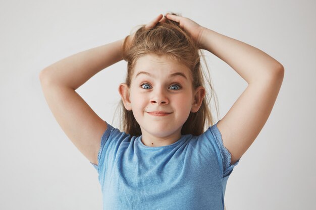 Ragazza bionda allegra divertente con gli occhi azzurri che tengono i capelli con le mani, con le sopracciglia sollevate e l'espressione sciocca del fronte