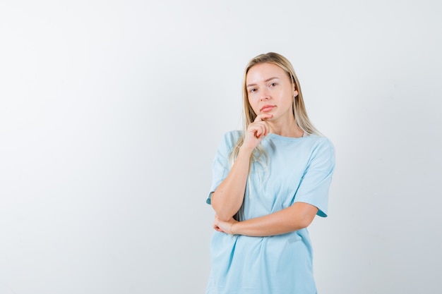 Ragazza bionda appoggiando il mento a portata di mano, in piedi nella posa di pensiero in maglietta blu e guardando pensieroso, vista frontale.