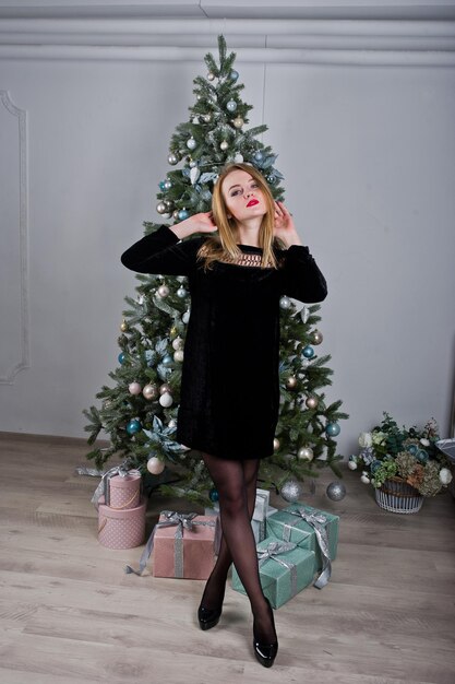 Ragazza bionda carina in abito nero contro l'albero di Capodanno con regali di Natale in studio Buone vacanze