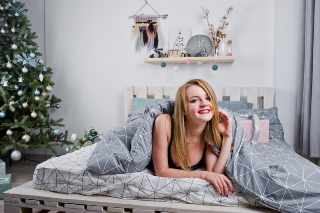 Ragazza bionda carina sul reggiseno nero sdraiato sul letto con scatole regalo contro l'albero di Capodanno in studio Buone vacanze