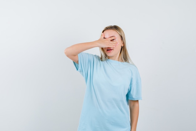 Ragazza bionda che copre gli occhi con la mano, guardando attraverso le dita in maglietta blu e guardando vergogna, vista frontale.