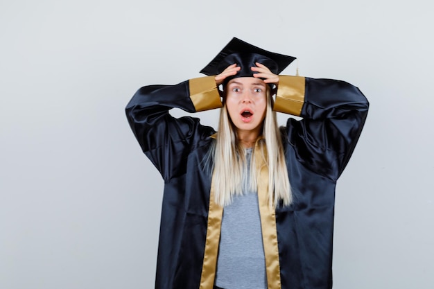 Ragazza bionda che copre la testa con le mani, aprendo la bocca in abito da laurea e berretto e sembra sorpresa.