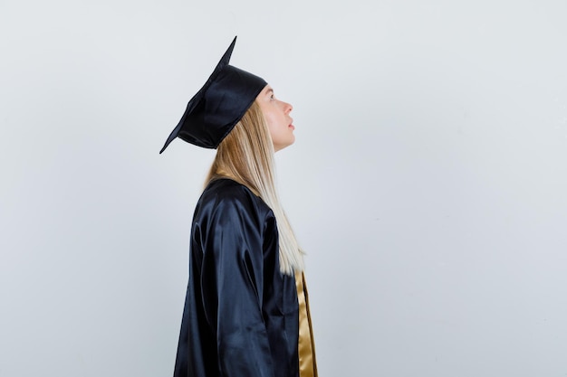 Ragazza bionda che guarda verso l'alto in uniforme laureata e sembra pensierosa.