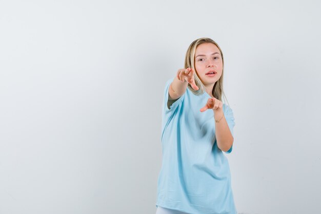 Ragazza bionda che indica alla macchina fotografica con il dito indice in maglietta blu e guardando fiducioso, vista frontale.