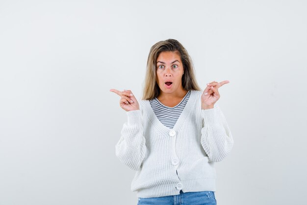 Ragazza bionda che indica direzioni opposte in maglietta a righe, cardigan bianco e pantaloni jeans e guardando sorpreso, vista frontale.