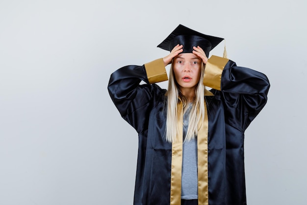 Ragazza bionda che mette le mani sulla fronte in abito di laurea e berretto e sembra sorpresa.