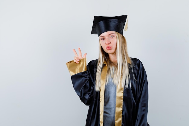 Ragazza bionda che mostra gesto di pace in abito e berretto da laurea e sembra carina