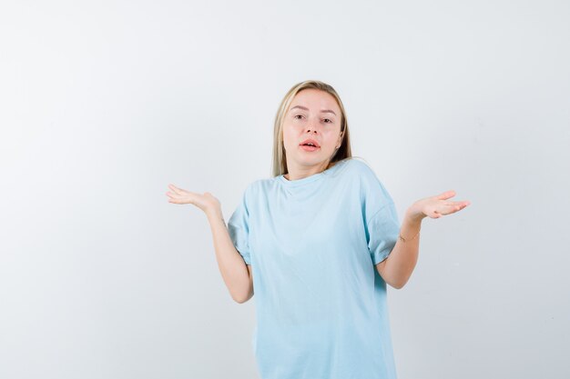 Ragazza bionda che mostra gesto impotente in maglietta blu e guardando perplesso, vista frontale.