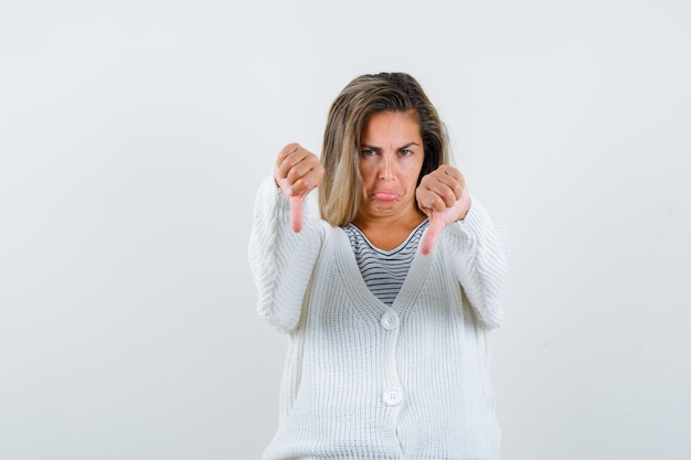 Ragazza bionda che mostra il doppio pollice verso il basso in maglietta a righe, cardigan bianco e pantaloni di jeans e sembra scontenta. vista frontale.