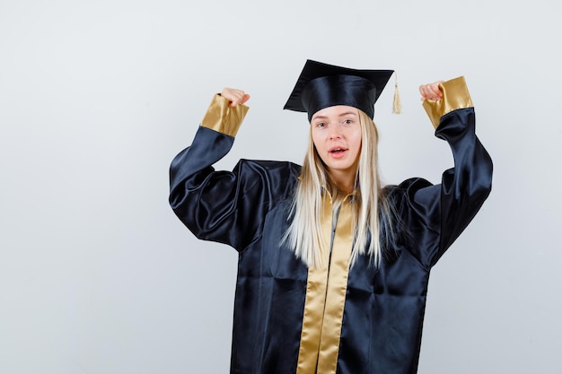 Ragazza bionda che mostra il gesto del vincitore in abito e berretto di laurea e sembra felice.