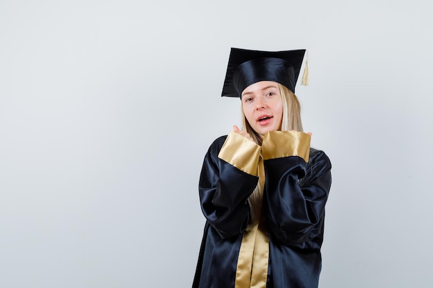 Ragazza bionda che mostra il gesto del vincitore in abito e berretto di laurea e sembra felice.