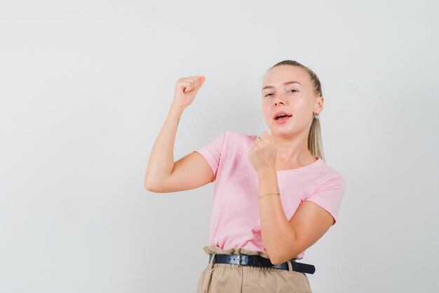 Ragazza bionda che mostra il gesto del vincitore in maglietta, pantaloni e che sembra fortunato, vista frontale.