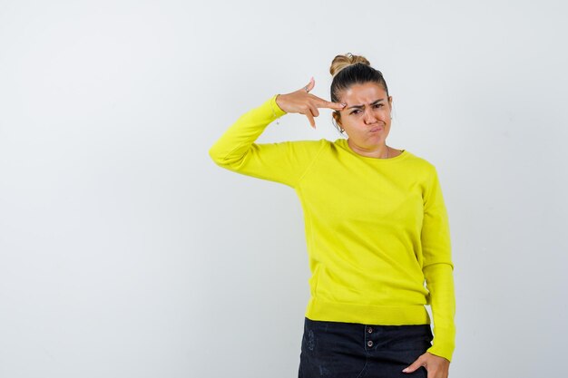 Ragazza bionda che mostra il gesto della pistola vicino alla testa, fa una smorfia con un maglione giallo e pantaloni neri e sembra infastidita