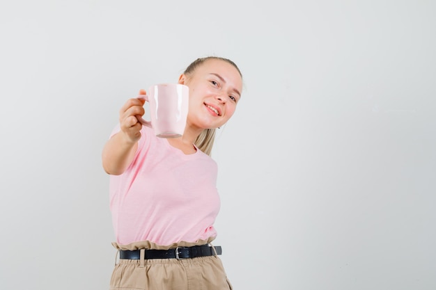 Ragazza bionda che mostra la tazza di bevanda in maglietta, pantaloni e che sembra felice. vista frontale.