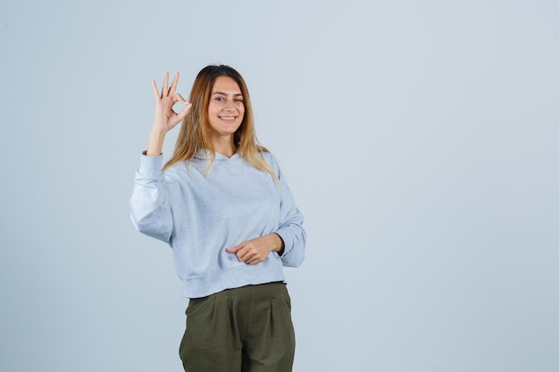 Ragazza bionda che mostra segno ok in felpa e pantaloni blu verde oliva e sembra raggiante, vista frontale.