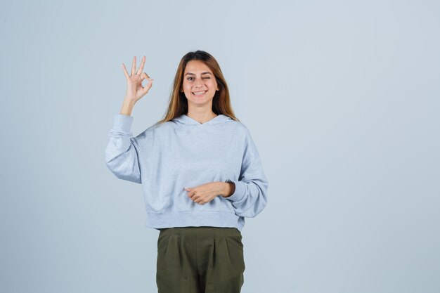 Ragazza bionda che mostra segno ok in felpa e pantaloni blu verde oliva e sembra raggiante, vista frontale.