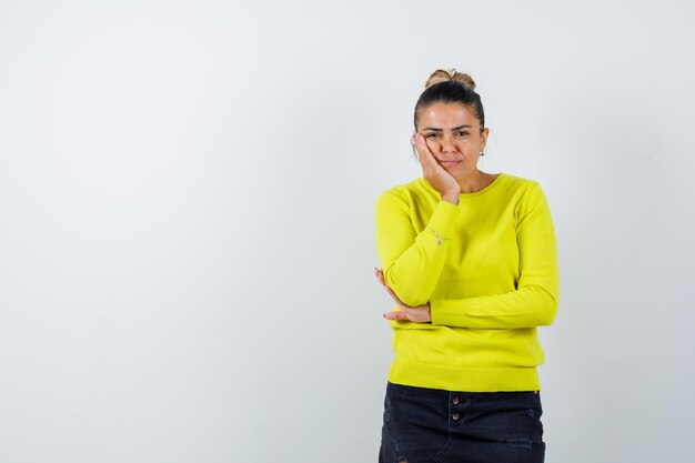 Ragazza bionda che si appoggia la guancia sul palmo mentre tiene la mano sul gomito con un maglione giallo e pantaloni neri e sembra pensierosa