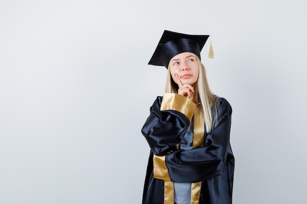 Ragazza bionda che sta nella posa di pensiero in abito e berretto di graduazione e che sembra pensierosa