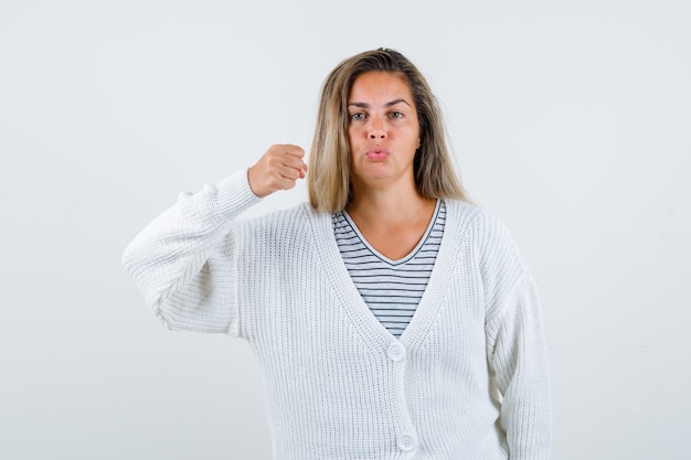 Ragazza bionda che stringe il pugno, labbra curve in t-shirt a righe, cardigan bianco e pantaloni di jeans e sembra sicura. vista frontale.