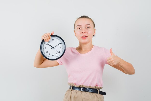 Ragazza bionda che tiene orologio da parete, mostrando il pollice in su in t-shirt, pantaloni e guardando allegro, vista frontale.