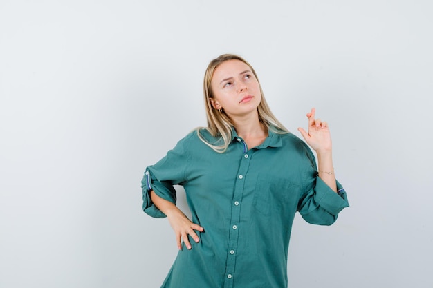 Ragazza bionda che tiene una mano sulla vita mentre tiene le dita incrociate in camicetta verde e sembra pensierosa