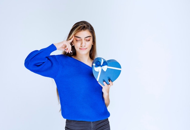 Ragazza bionda che tiene una scatola regalo blu a forma di cuore e pensa.