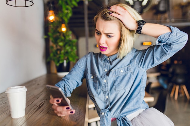 Ragazza bionda con labbra rosa brillante seduto in una caffetteria a bere caffè e guardando il suo smartphone. Sembra sorpresa e gioca con i suoi capelli. Indossa una camicia di jeans blu