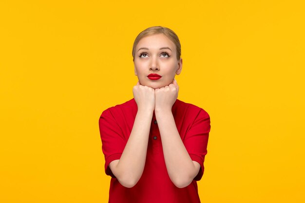 Ragazza bionda di giorno della camicia rossa che osserva in su i pugni sotto il mento in una camicia rossa su una priorità bassa gialla