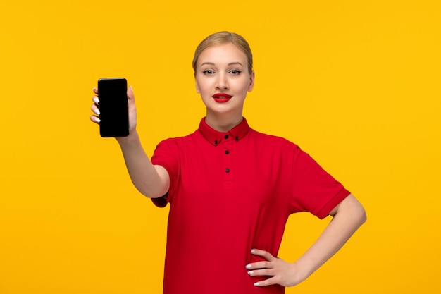 Ragazza bionda di giorno della camicia rossa che tiene il telefono nella mano in una camicia rossa su una priorità bassa gialla