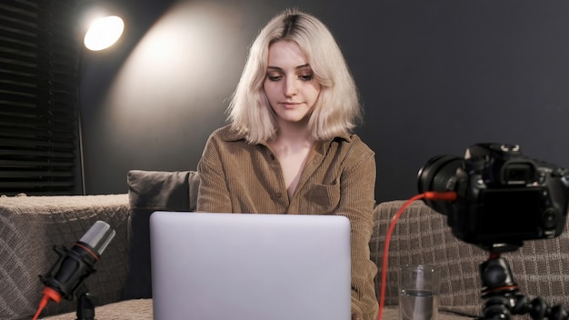 Ragazza bionda giovane creatore di contenuti con le cuffie che lavorano al suo computer portatile sul tavolo con la fotocamera su un treppiede