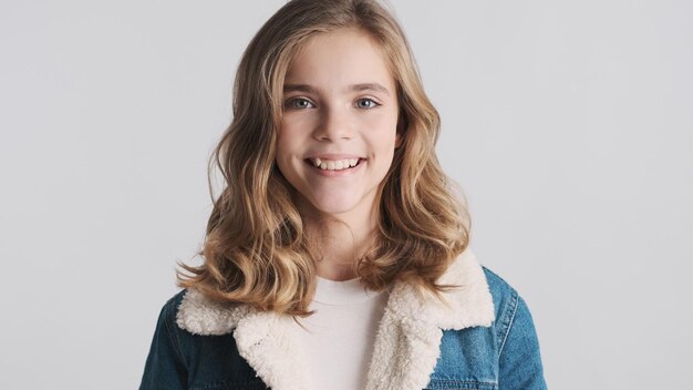 Ragazza bionda graziosa dell'adolescente con i capelli ondulati che sorride sulla macchina fotografica che sembra allegra isolata su fondo bianco. Emozioni positive