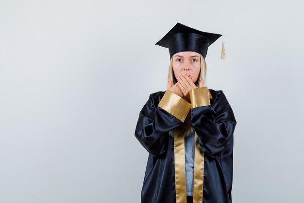 Ragazza bionda in abito da laurea e berretto che copre la bocca con le mani e sembra sorpresa