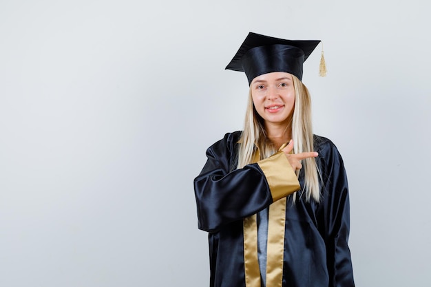 Ragazza bionda in abito da laurea e berretto che punta a destra con il dito indice e sembra carina