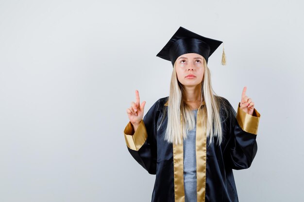 Ragazza bionda in abito da laurea e berretto che punta con l'indice e sembra carina