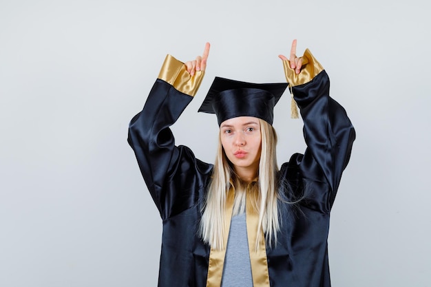 Ragazza bionda in abito da laurea e berretto che punta con l'indice e sembra carina