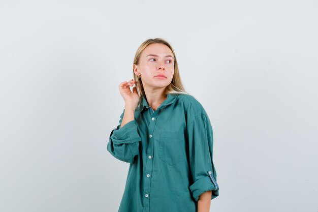 Ragazza bionda in camicetta verde che guarda lontano mentre posa in macchina fotografica e sembra incantevole