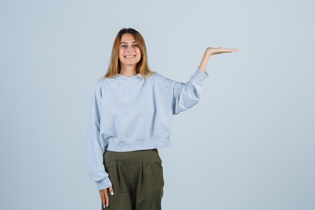 Ragazza bionda in felpa blu verde oliva e pantaloni che allungano le mani mentre tiene qualcosa e sembra raggiante, vista frontale.