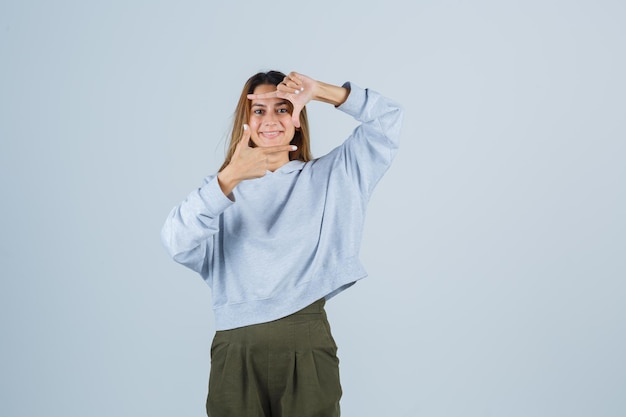 Ragazza bionda in felpa blu verde oliva e pantaloni che mostra il gesto della fotocamera e sembra raggiante, vista frontale.