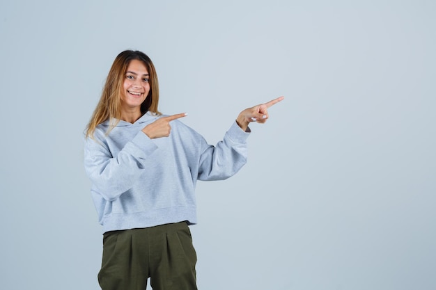 Ragazza bionda in felpa blu verde oliva e pantaloni che puntano a destra con le dita indice e sembra affascinante, vista frontale.