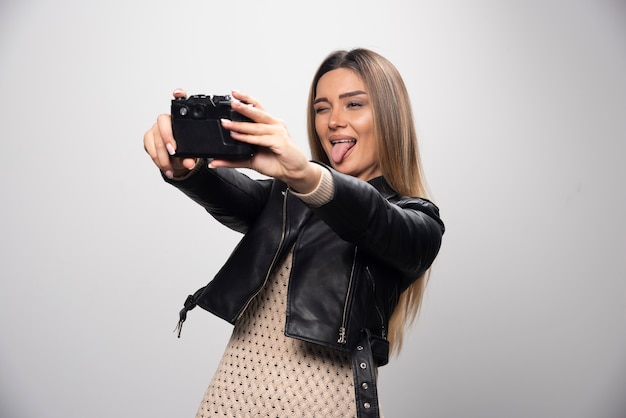 Ragazza bionda in giacca di pelle nera prendendo i suoi selfie con una macchina fotografica.