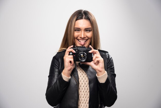 Ragazza bionda in giacca di pelle nera prendendo i suoi selfie con una macchina fotografica.