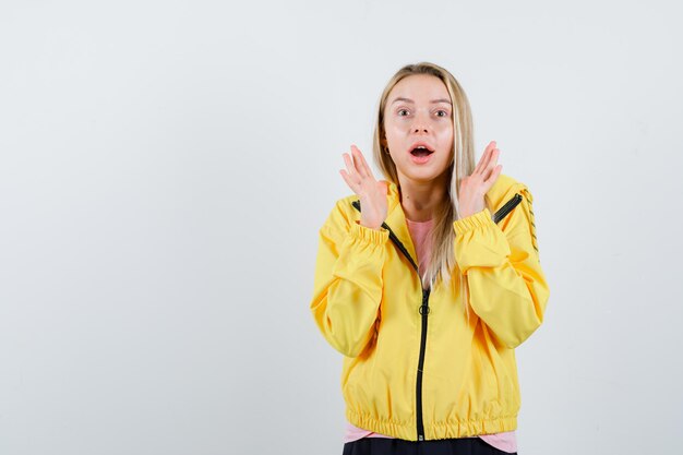 Ragazza bionda in giacca gialla che alza le mani, apre la bocca e sembra sorpresa