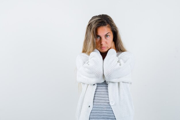 Ragazza bionda in maglietta a righe, cardigan bianco e pantaloni jeans che mette le mani sul collo e sembra seducente, vista frontale.