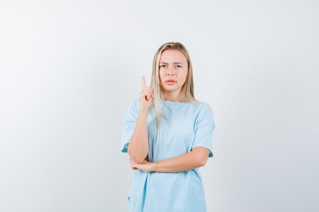 Ragazza bionda in maglietta blu che alza il dito indice nel gesto di eureka mentre si tiene la mano sotto il gomito e sembra sensibile, vista frontale.